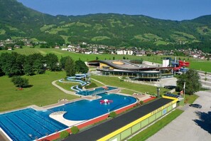 Ein Schwimmbad mit Wasserrutsche, ein Gebäude mit gewölbter Dachform und eine gebirgige Landschaft im Hintergrund.