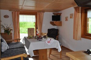 Ein gemütliches Zimmer mit einer Couch, einem Tisch mit weißer Tischdecke, einem an einer Holzablage montierten Fernseher und einem Fenster mit Vorhängen.
