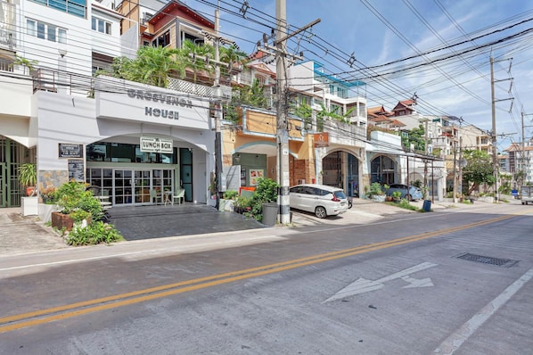 Vista da rua com uma casa para alugar, placa indicando serviço de refeições e carros estacionados.