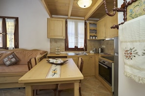 A cozy kitchen with wooden cabinets, a dining table with chairs, a built-in oven, and a window with curtains. - Kaminos Boutique Hotel (Symi)