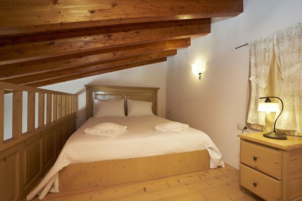 A bedroom with a wooden bed, a nightstand, a lamp, and a window with curtains. - Kaminos Boutique Hotel (Symi)