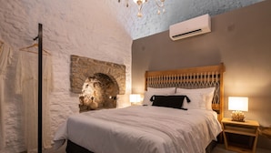 A bedroom with a stone wall, a wooden bed frame, a hanging light fixture, and an air conditioning unit. - Kaminos Boutique Hotel (Symi)