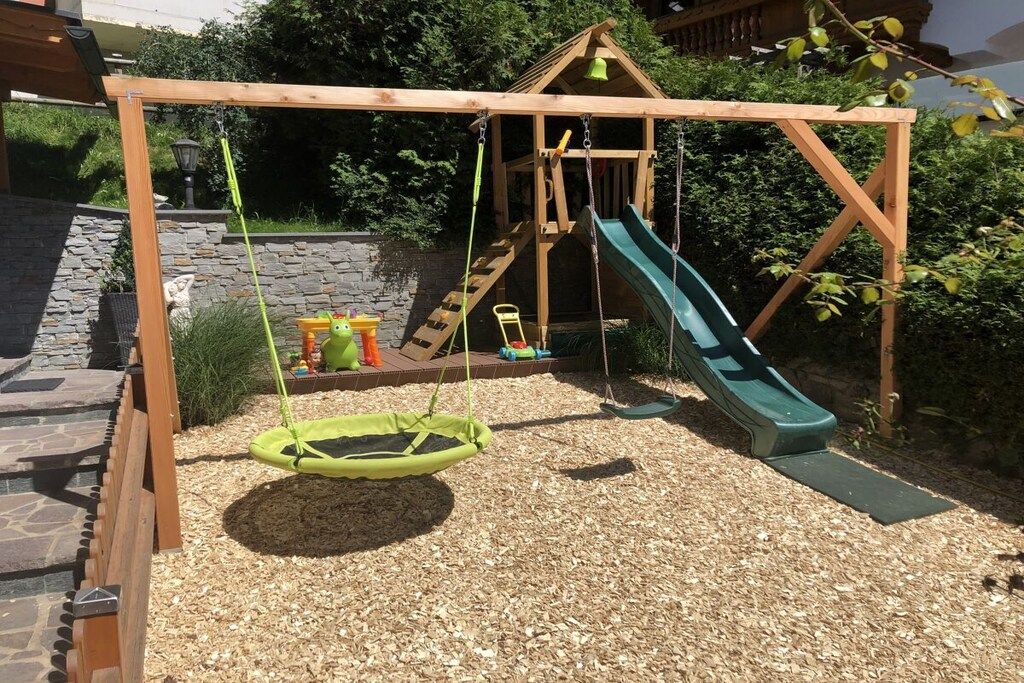 A wooden swing set with a slide, surrounded by a gravel area and greenery.