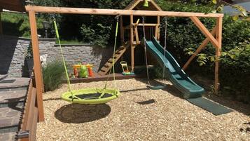 A wooden swing set with a slide, surrounded by a gravel area and greenery.