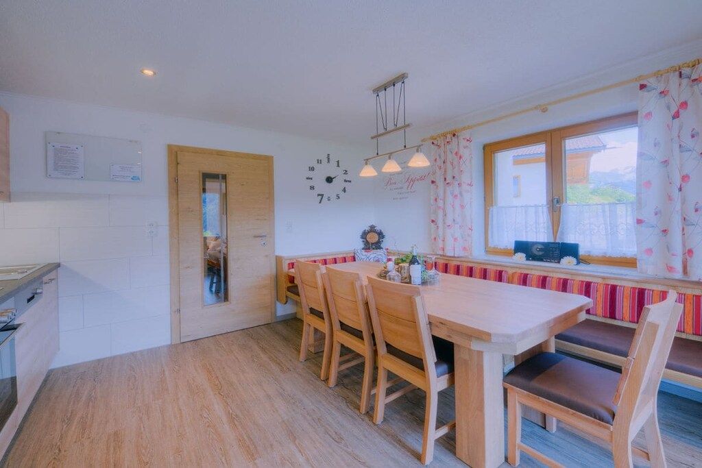 A kitchen with a wooden table and chairs, a window with curtains, and a clock on the wall.