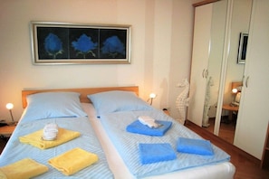 Een hotelkamer met twee bedden, elk met een blauwe plaid en kussens, een lijst met daarin een schilderij van blauwe bloemen aan de muur, en een kledingkast met spiegel.