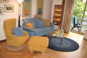 A living room with a blue sofa, yellow armchair, and ottoman, a glass coffee table, and a view of the outdoors. - 08 Heringsdorf Lake Castle (Heringsdorf)
