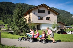 A family with children on a small vehicle in front of a house with a mountain backdrop. - in the Bergquell house with horse stable (Au, Bregenzerwald)