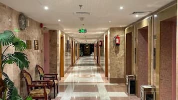 A hotel corridor with a checkered floor, chairs, potted plants, and doors leading to rooms.