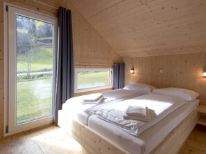 Ein Schlafzimmer einer Holzhütte mit einem großen Bett, zwei Fenstern und Blick auf eine grüne Landschaft.