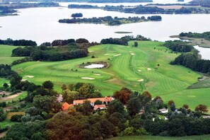 Een golfbaan met een waterpartij op de achtergrond, omgeven door bomen en een gebouw met een rood dak.