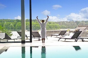 Eine Person in einem weißen Bademantel steht an einem Pool mit Liegestühlen und Blick auf eine bewaldete Landschaft.