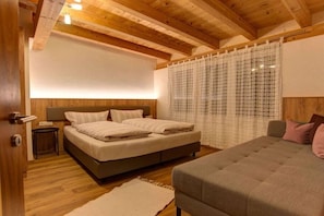 A bedroom with a bed, a sofa, wooden flooring, a headboard, and a window with curtains. - Holiday apartment with 1 bedroom (Pettneu am Arlberg)