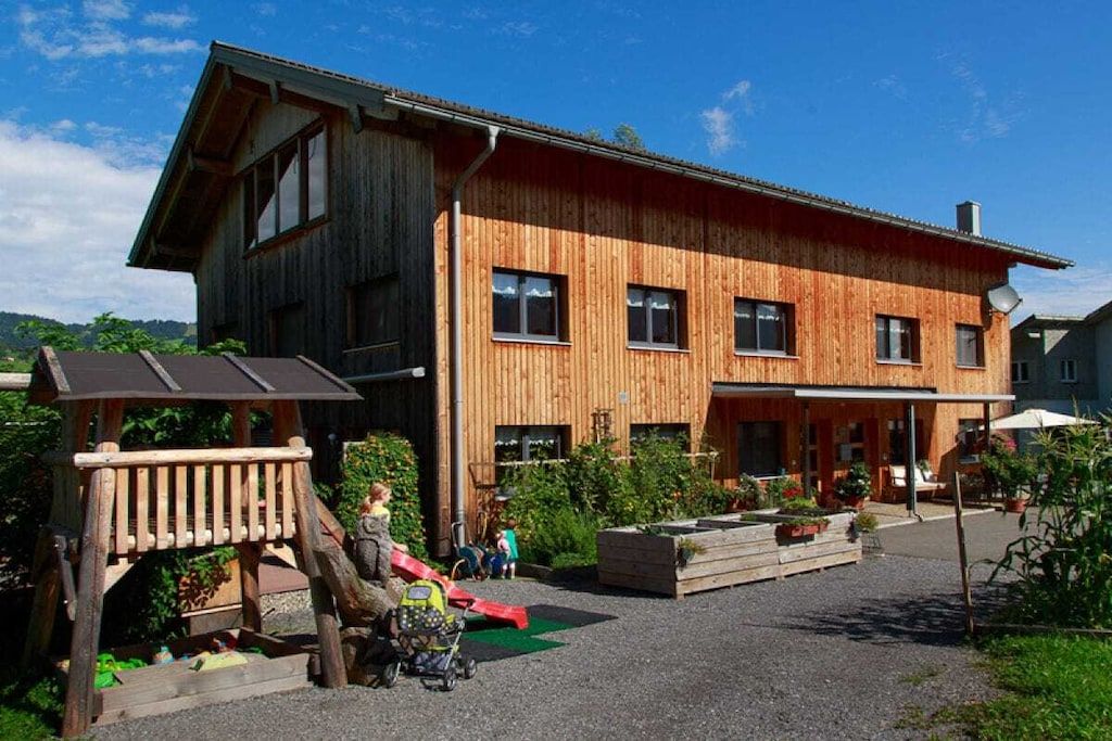 Un bâtiment en bois doté d’un toit en pente, d’un espace extérieur couvert avec des sièges et d’une aire de jeux.