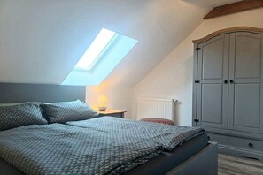 Ein Schlafzimmer mit geneigter Decke, einem Oberlicht, einem Bett mit gemusterten Betttextilien, einem Nachttisch mit Lampe und einem großen Kleiderschrank.
