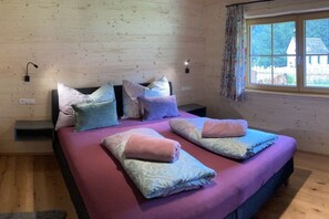 A bedroom with a bed, pillows, a nightstand, a window with curtains, and a view of a house. - Nature Chalet Pitztal (Wenns)