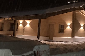 A building with a covered outdoor area, snow on the ground, and lights illuminating the structure. - Nature Chalet Pitztal (Wenns)