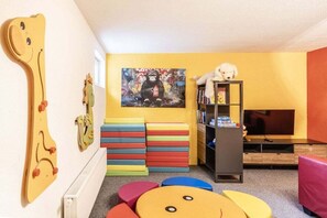 Ein Zimmer mit Spielbereich, inklusive bunter Bausteine und einer Spielgitarre, einem Bücherregal mit Büchern und einem Teddybär sowie einem Bild an der Wand.
