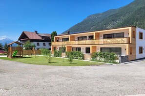 Ein zweistöckiges Gebäude mit einem Holzbalkon, einem Spielbereich für Kinder inklusive Rutsche und einem Berg im Hintergrund.