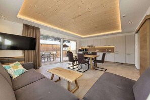 A modern living room with a flat-screen TV, a sofa, a dining table with chairs, and a kitchen area with cabinets. - 80m² Guesthouse Sonne (Bichlbach)