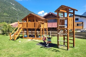 A wooden play structure with stairs and a swing set in a grassy area with mountains in the background. - 80m² Guesthouse Sonne (Bichlbach)