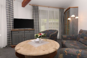 Ein Zimmer mit Fernseher, einem runden Holztisch mit Blumenvase, einer Couch und Vorhängen.