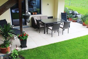 Eine Terrasse im Freien mit Esstisch und Stühlen, umgeben von Blumentöpfen und einer Rasenfläche.