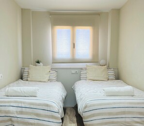 A room with two beds, each with striped bedding and pillows, a window with curtains, and a small plant on the left side. - Elegant Coastal Apartment with Marina & Beach View (Port Saplaya)