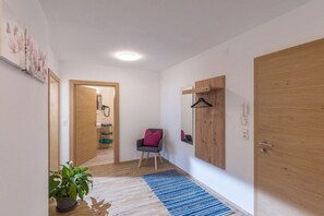 A hallway with a wooden floor, a blue rug, a potted plant, a chair with a pink cushion, and a wooden door. - Holiday apartment with 1 bedroom (Ramsau)