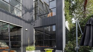 A modern outdoor seating area with a round table and white wireframe chairs, surrounded by a building with large windows and vertical siding. - Melbourne City Apartments Luxury Loft House with rooftop (Southbank)