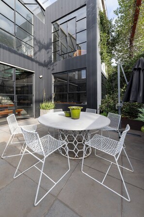 Uma área de estar moderna ao ar livre, com uma mesa redonda e cadeiras de estrutura branca, cercada por um edifício com janelas amplas e revestimento vertical.