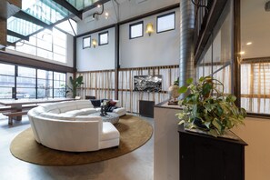 Uma sala ampla com um sofá modular branco, uma mesa de centro redonda e uma planta grande em vaso.