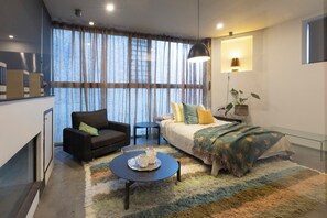 Quarto moderno com sofá, cama com manta colorida, mesa de centro com bandeja e uma janela grande com cortinas.