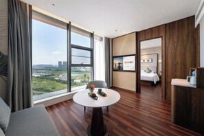 Een hotelkamer met een groot raam, een ronde tafel met een schaal fruit, een bed en een houten vloer.