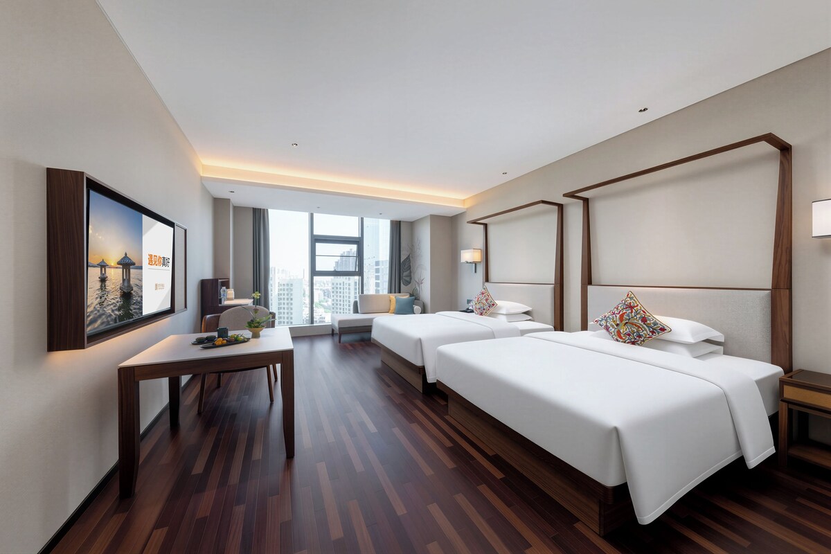 A hotel room with two beds, a desk, and a large window.