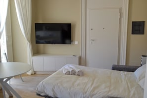 Hotellihuone, jossa on sänky, seinälle asennettu televisio, ovi ja pieni pöytä.