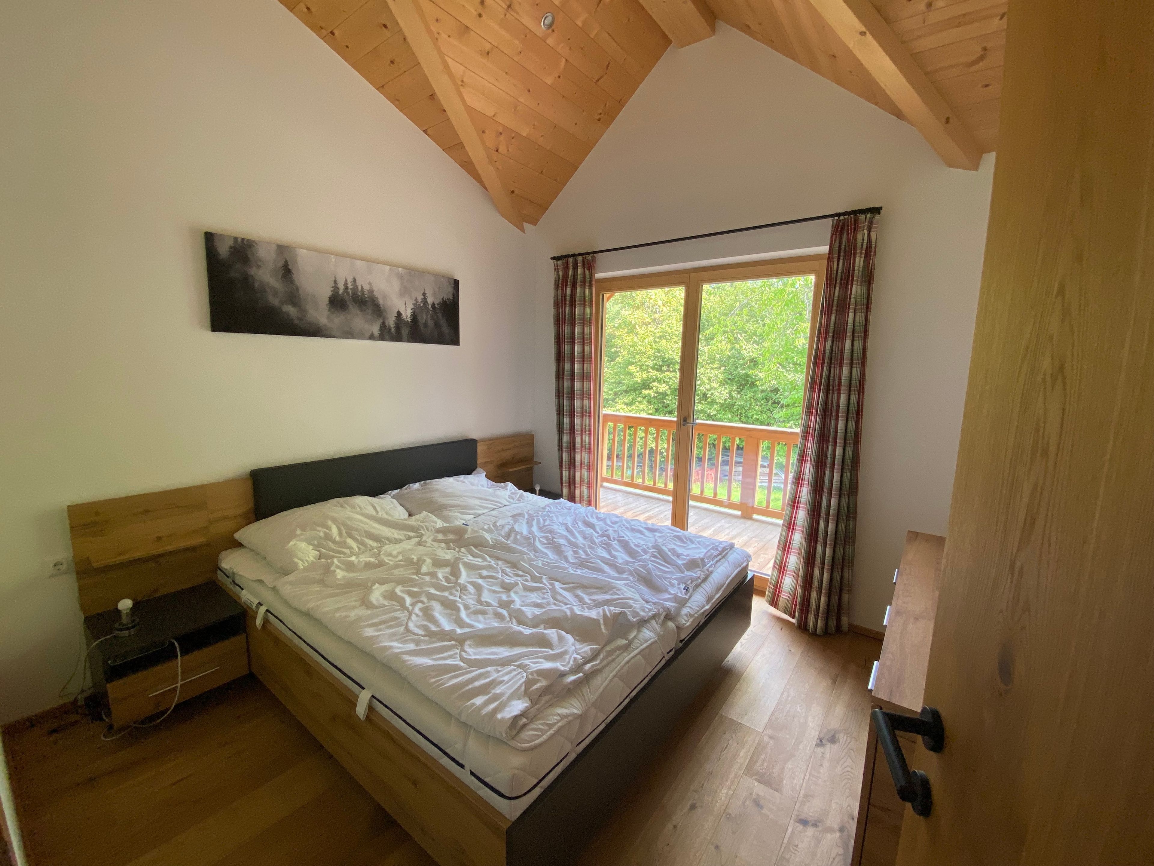 A bedroom with a wooden ceiling, a bed, a nightstand, a painting, and a balcony with curtains.