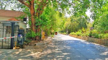 A tree-lined street with a gated entrance, a parked auto-rickshaw, and a clear sky.