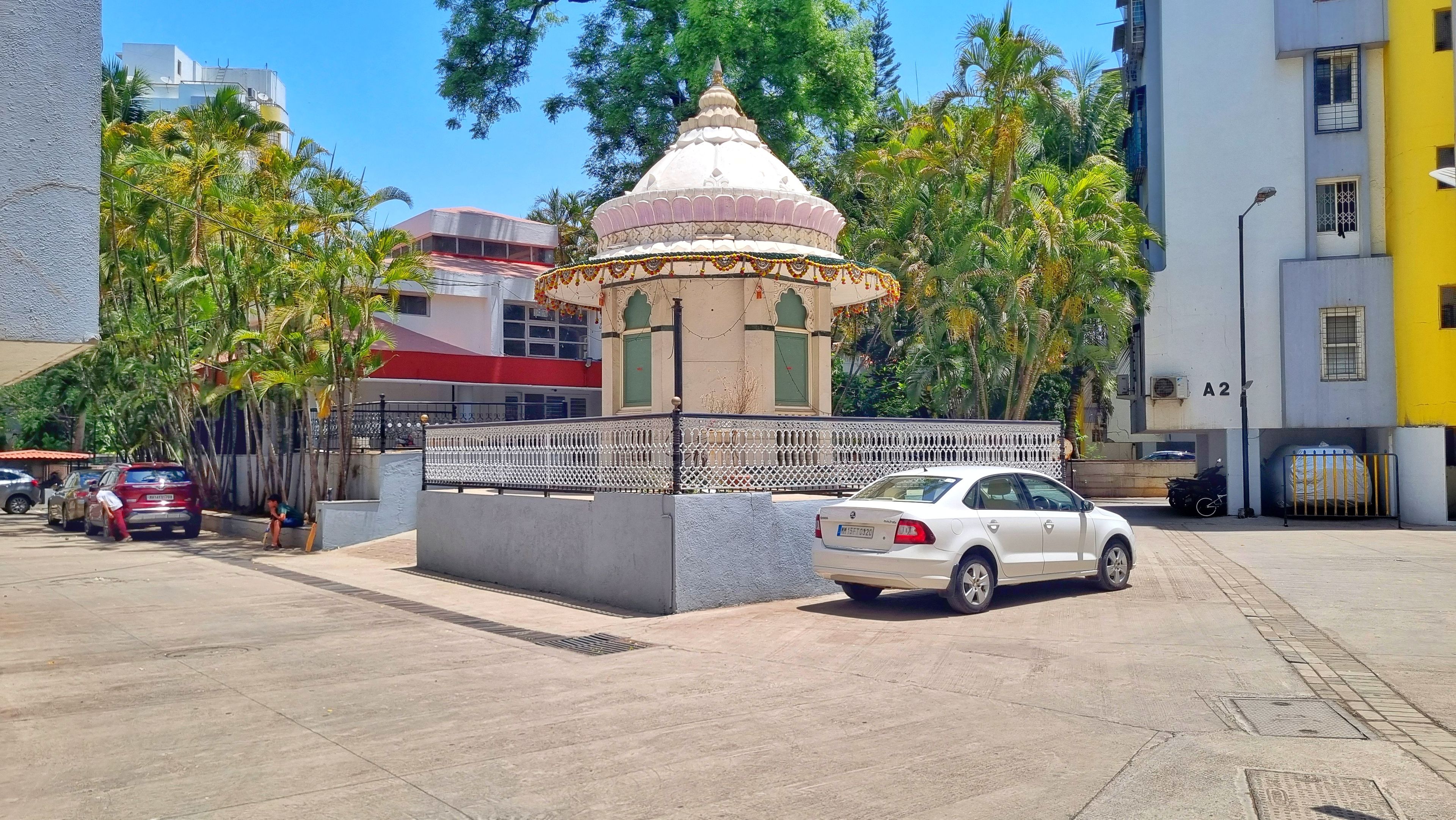 A white car parked in front of a small, white building with a dome roof, surrounded by palm trees and a fence.