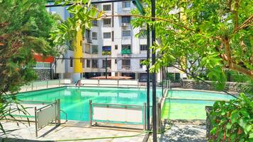 A swimming pool with a surrounding deck, a multi-story building in the background, and lush greenery.