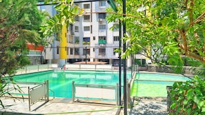 Una piscina con un deck circundante, un edificio de varios pisos al fondo y abundante follaje.