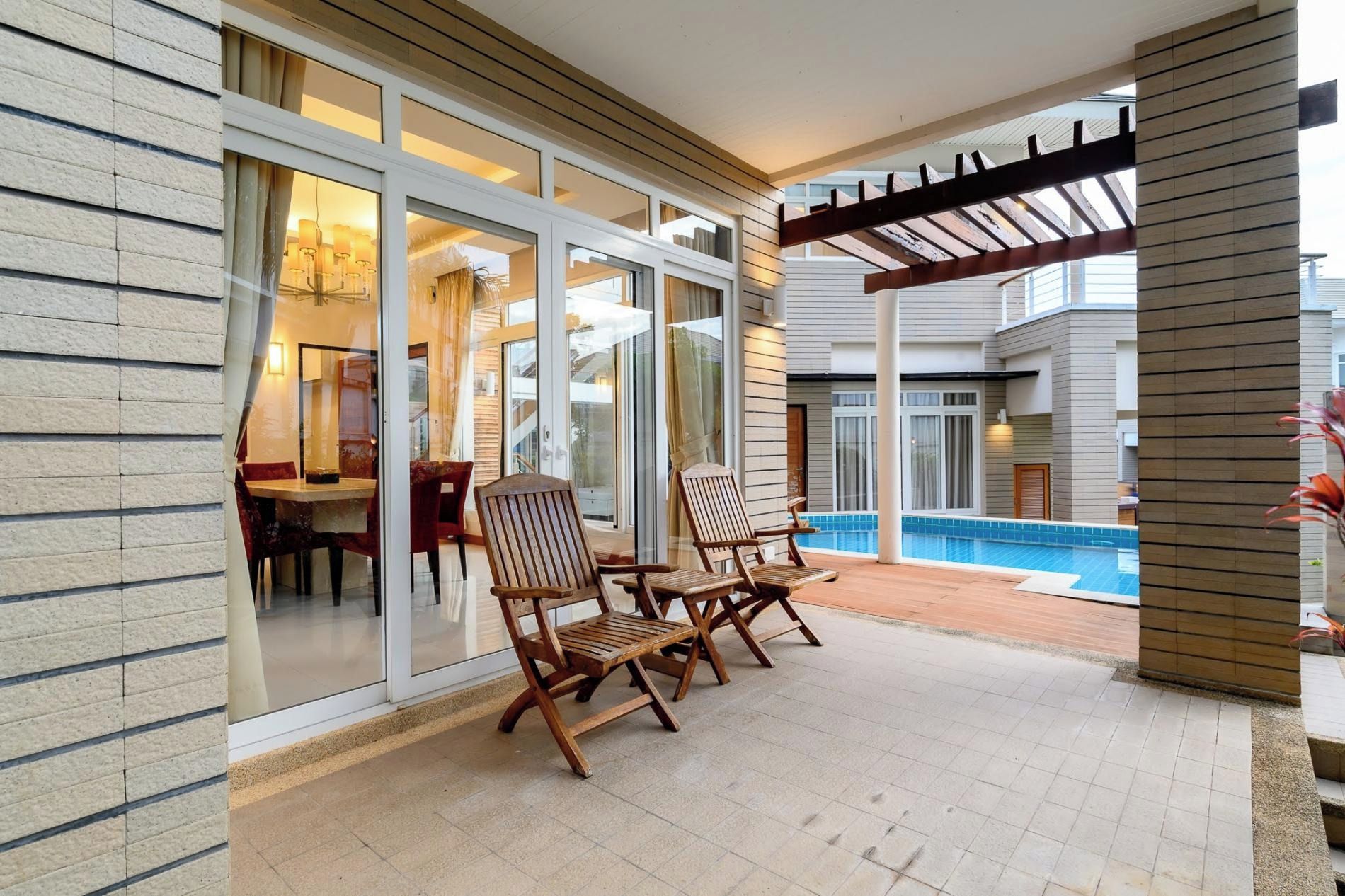 A covered outdoor area with a pool, wooden chairs, and a dining table.