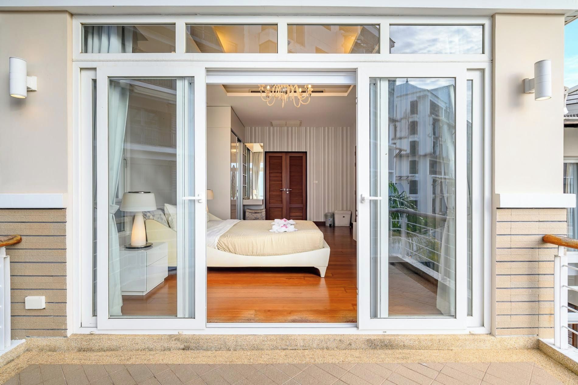 A modern bedroom with a large sliding glass door, a bed with a beige duvet, a nightstand with a lamp, and a chandelier.