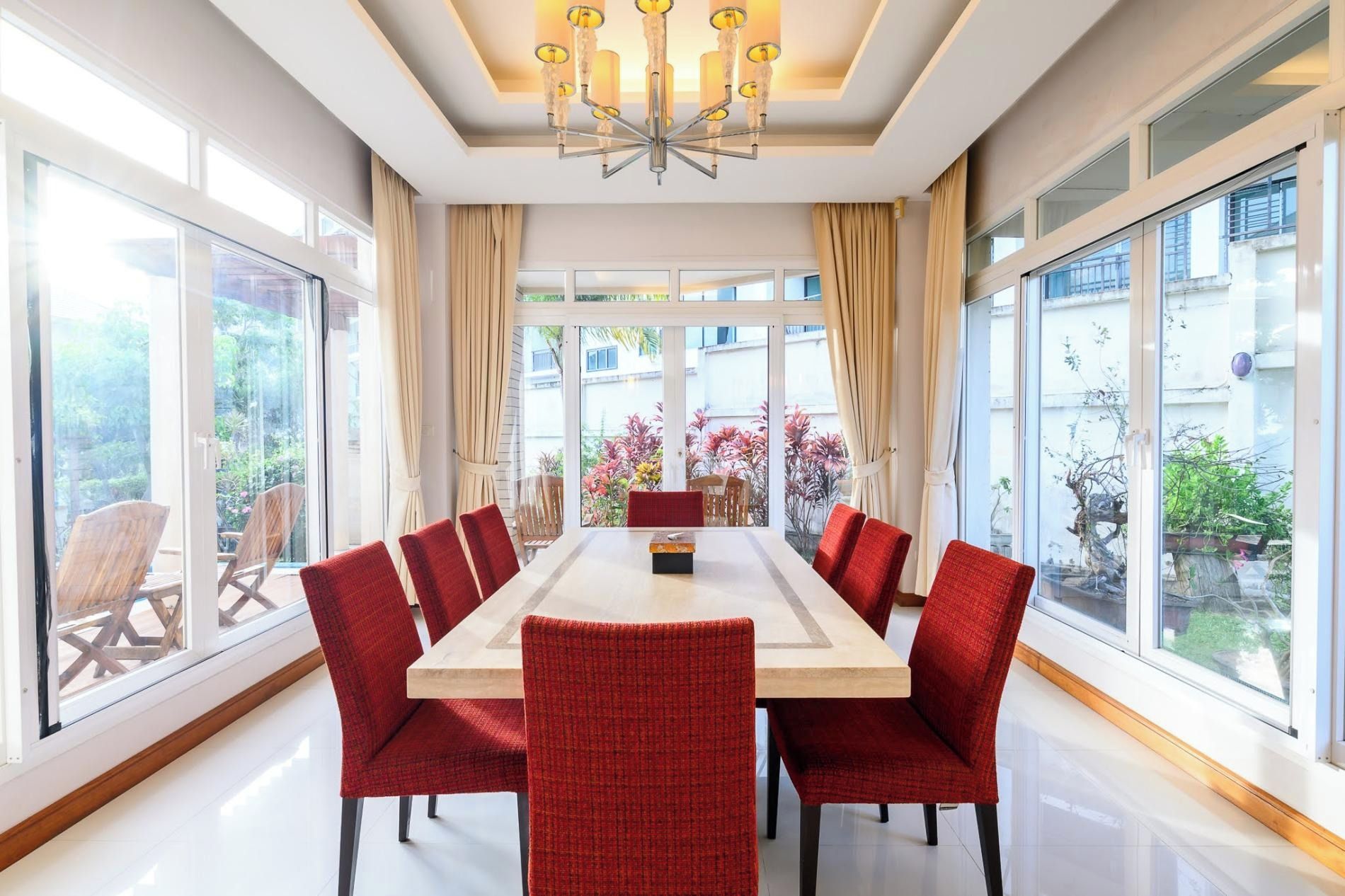 Un comedor con una mesa larga y sillas rojas, ventanas amplias y un candelabro.