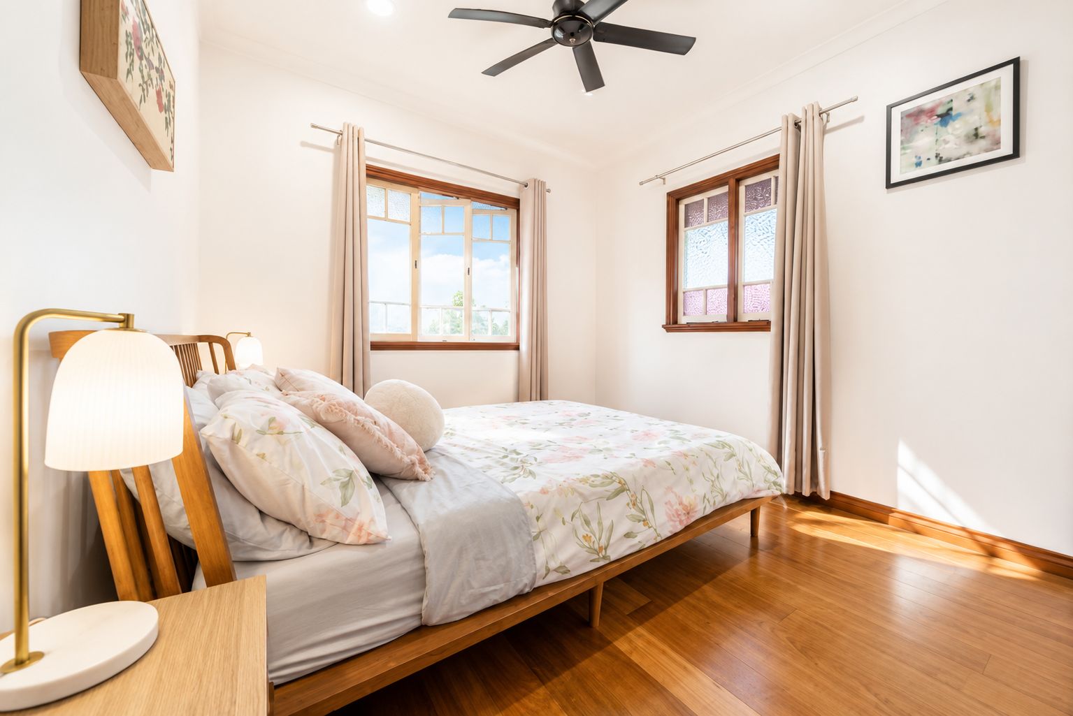 A bedroom with a bed, a bedside table, a lamp, a ceiling fan, and two windows with curtains.