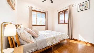 A bedroom with a bed, a bedside table, a lamp, a ceiling fan, and two windows with curtains.