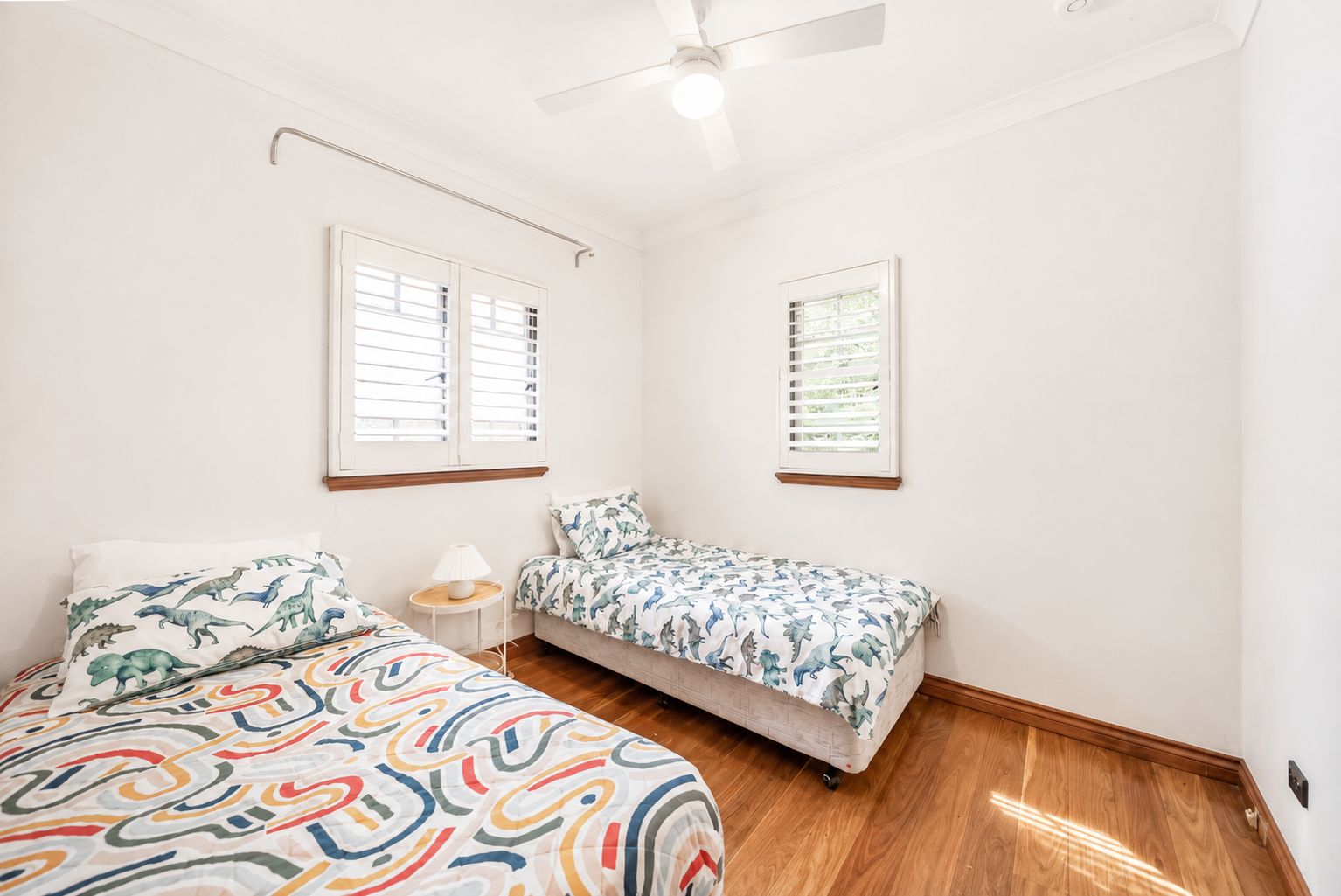 A bedroom with a bed, a small sofa, two windows with blinds, a ceiling fan, and a wooden floor.
