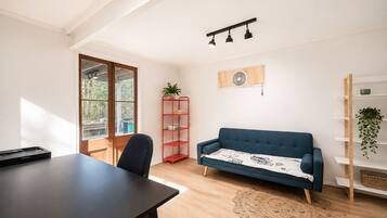 A room with a dark blue sofa, a red shelving unit, a wooden floor, and a large window with a view of trees.