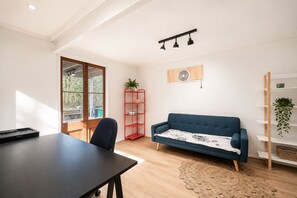 A room with a dark blue sofa, a red shelving unit, a wooden floor, and a large window with a view of trees.