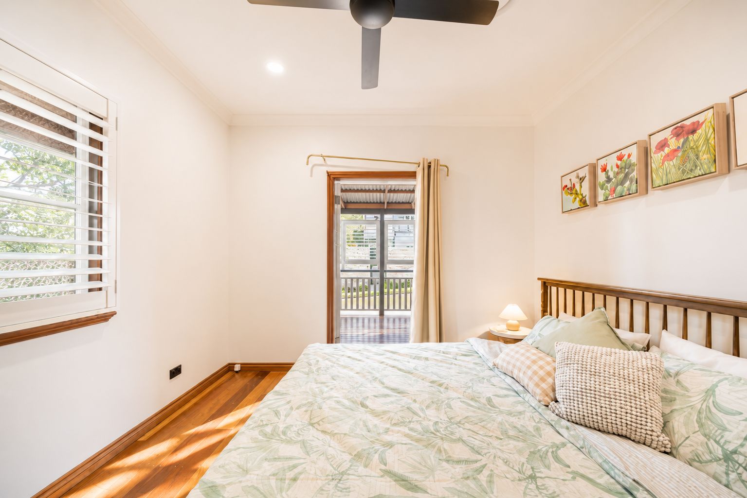 A bedroom with a bed, a ceiling fan, a window with blinds, a door leading to an outdoor area, and artwork on the wall.